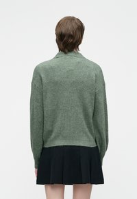 Persona con cabello corto castaño, vistiendo un suéter verde con textura acanalada y una falda negra con pliegues, de pie frente a un fondo blanco liso.