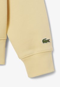 Manga de sudadera amarilla, puño acanalado, textura suave, con un logo de cocodrilo bordado en verde en la parte superior del brazo.