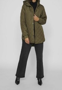 Olivgrön quiltad jacka med huva, försedd med tryckknappar och elastiska muddar, kombinerad med svarta utsvängda jeans och spetsiga svarta stövlar.