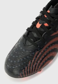 Μαύρο ποδοσφαιρικό παπούτσι με υφή στην επιφάνεια, πορτοκαλί διαγώνιες γραμμές, μικρά μοτίβα μπάλας ποδοσφαίρου και μαύρα κορδόνια δεμένα στην κορυφή.