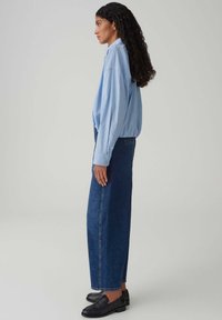 Lichtblauw gestreept oversized overhemd, lange donkerblauwe recht uitgesneden spijkerbroek en zwarte loafers, zijaanzicht tegen een neutrale achtergrond.