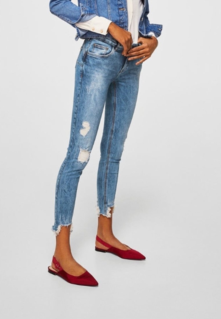 Personne portant un jean skinny bleu clair déchiré avec des ourlets effilochés, des chaussures plates rouges à bride arrière pointues, et une veste en jean sur un t-shirt blanc.