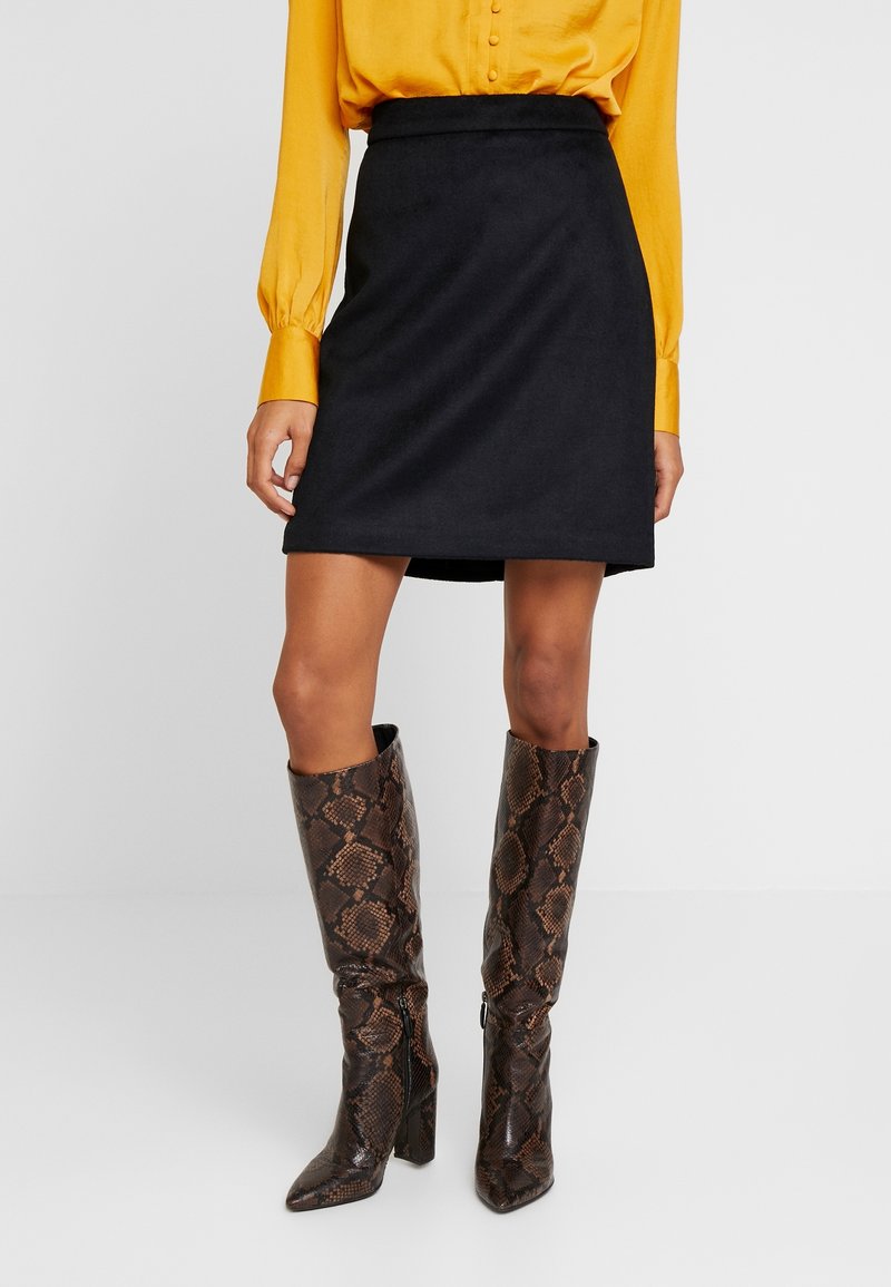 Femme portant une jupe noire mi-longue, une chemise jaune boutonnée et des bottes marron à motif peau de serpent jusqu'aux genoux.