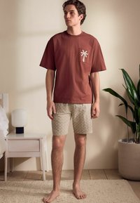 Czerwono-rdzawy bawełniany t-shirt z wzorem palmy na kieszeni, w połączeniu z beżowymi szortami w wzory. Na boso, w neutralnym otoczeniu pokoju.