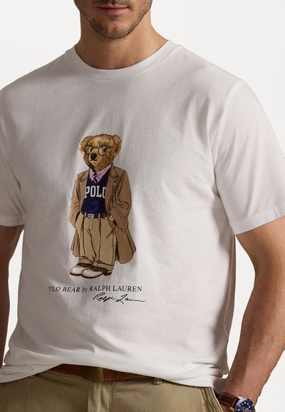 Bela majica z grafiko medveda v rjavem plašču, vijoličastem puloverju, bež hlačah in očalih, s tekstom "POLO BEAR by RALPH LAUREN".