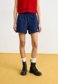 Eine Person in navyfarbenen Adidas-Shorts mit floralen seitlichen Streifen, einem roten gestrickten ärmellosen Oberteil, weißen Socken und schwarzen Schnürstiefeln steht auf einem gelben Boden.