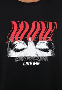 Svart t-shirt med en grafisk design av stängda ögon, med röd text "INGEN" ovanför och vit text "SER SPELET SOM JAG" nedan.