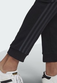 Underbenen bär svarta byxor med mörka ränder och vita Adidas Superstar-sneakers med svarta och guldiga detaljer mot en ljus bakgrund.