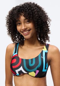 Γυναίκα με χαμόγελο και σγουρά μαλλιά, φοράει πολύχρωμο αθλητικό σουτιέν με γεωμετρικά σχέδια, σε απλό φωτεινό φόντο.