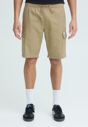 Mann trägt beige Cargoshorts mit Seitentaschen, weiße Sportsocken und schwarze Turnschuhe, steht vor einem schlichten hellen Hintergrund.