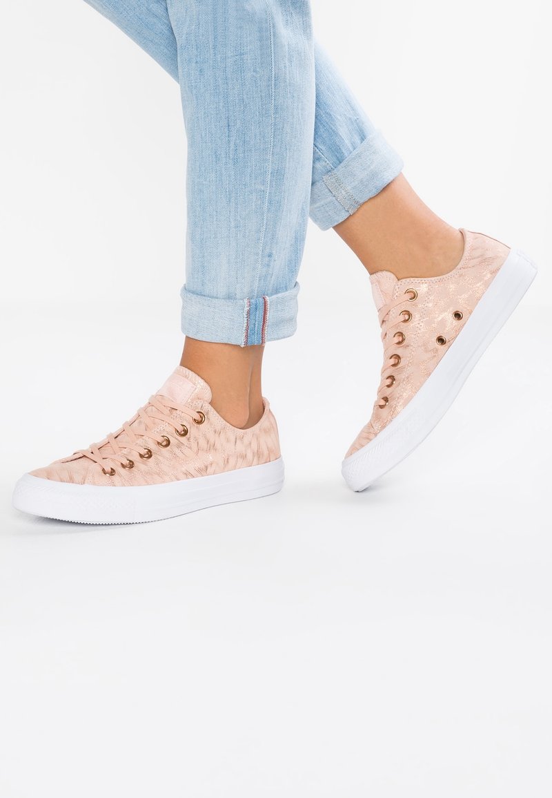 Personne portant un jean bleu clair retroussé et des baskets roses claires à motifs avec des semelles blanches, debout sur un sol blanc.
