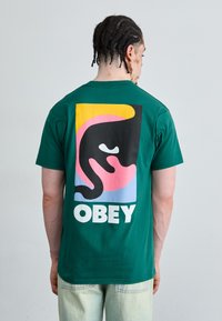 Zöld pamut póló, nagy színes grafikával a hátsó részén, absztrakt formákkal és vastag, fehér betűkkel írva: 'OBEY'.