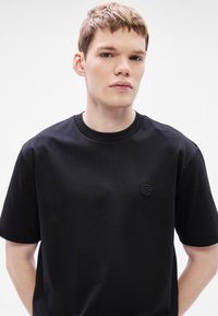 Fekete pamut rövid ujjú póló, kerek nyakkivágással és a mellkason egy kis dombornyomott logóval. Simább textúra, enyhén laza szabás.