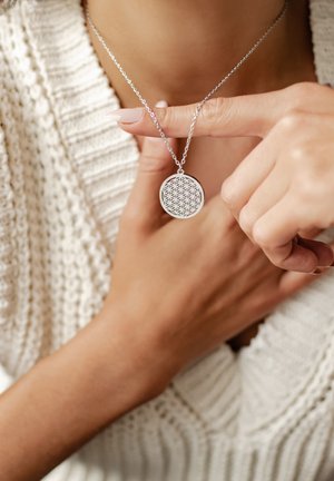 Srebrny wisiorek o geometrycznym wzorze, okrągły kształt, zawieszony na cienkim łańcuszku. Modelka nosi teksturowany biały sweter w splotach.