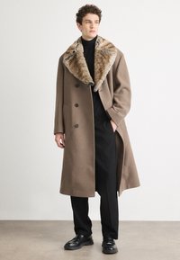 Abrigo de lana marrón con un cuello de piel sintética, diseño de doble botonadura, grandes botones y bolsillos laterales, combinado con pantalones y zapatos negros.