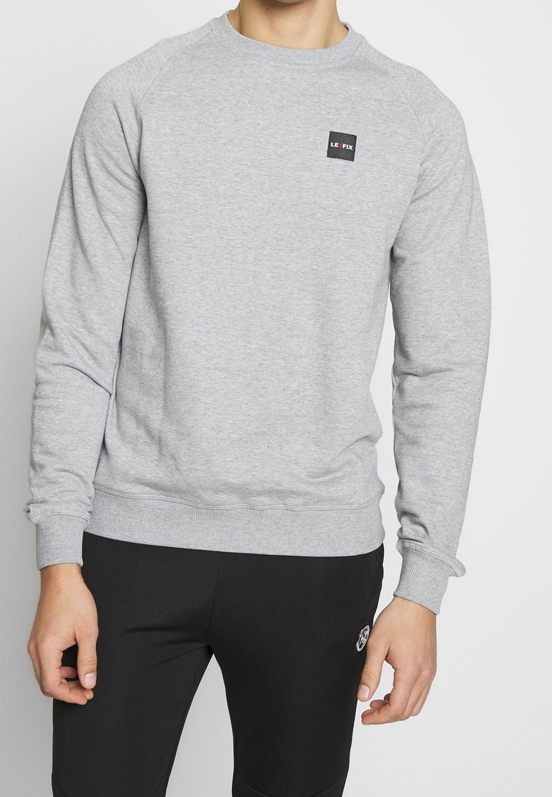 Homme portant un sweat-shirt à manches longues gris clair avec un petit logo noir sur la poitrine et un pantalon noir avec un petit emblème rond sur la cuisse.
