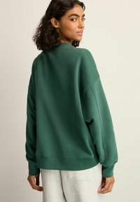 Groene sweatshirt met een ronde hals, losse pasvorm, geribde manchetten en een gladde textuur. Draagt met lichtgrijze joggers, gezien van achteren.