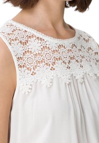 Blusa blanca suelta con un empaque de encaje floral, que presenta delicados bordados y acentos texturizados en el escote y el dobladillo. Tejido suave y ligero.