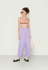 Top multicolor cortado com padrão em zig-zag, combinado com calças lilás de cintura alta e sandálias brancas com solas pretas. Os acessórios incluem óculos de sol e uma bolsa bege.