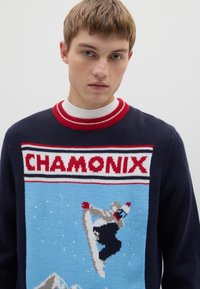 Marineblauer Strickpullover mit einem Snowboard-Design aus Chamonix, einem rot-weiß gestreiften Kragen und einem bunten Grafikmuster auf der Vorderseite.