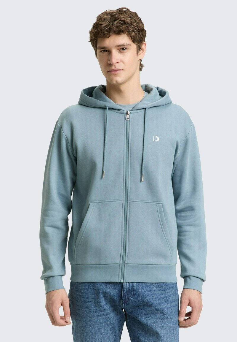 Giovane con capelli ricci che indossa una felpa con cappuccio azzurra con zip e jeans blu, in piedi contro uno sfondo chiaro e uniforme.