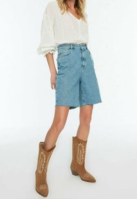 Mujer con blusa blanca de manga larga, pantalones cortos de mezclilla a la rodilla de talle alto y botas de vaquero marrones bordadas.