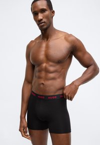 Melni bokseršorti no elastīga auduma ar sarkanu "HUGO" logotipu uz jostasvietas. Dizains ir pieguļošs un gluds, izceļot ķermeņa kontūras.
