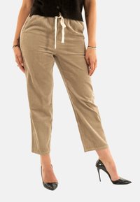 Femme portant un pantalon en velours côtelé beige avec un cordon blanc et des chaussures à talons hauts noires, se tenant les mains détendues sur les côtés.