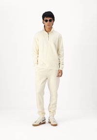 Kremast dukserica s polu-rajmericom, u kombinaciji s odgovarajućim jogger hlačama. Outfit se ističe mekim tekstilom i elastičnim manžetama.