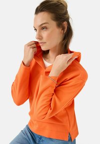 Oranje pullover hoodie met een relaxte pasvorm, hoge hals en zijsplitten. Gemaakt van een gladde stof, met zichtbare stikseldetails en lange mouwen.