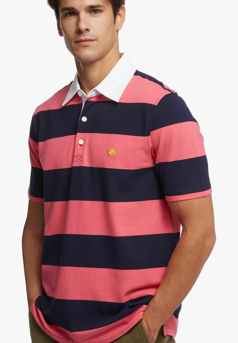 Růžovo-námořnický pruhovaný polo tričko z bavlny, které má bílý límec a malé žluté logo na hrudi. Krátké rukávy.