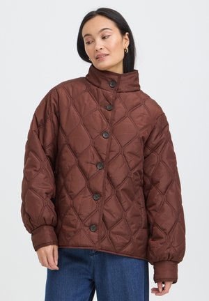 Femme portant une veste matelassée marron avec de gros boutons et un col montant, associée à un jean bleu, sur un fond clair uni.