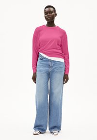 Rosa Langarmshirt über einem weißen Top, kombiniert mit hoch taillierten, lockeren hellblauen Jeans und silbernen Schuhen. Entspannte Passform und lässiger Stil.