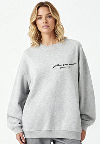 Femme aux cheveux blonds portant un sweat-shirt oversize gris clair avec l'inscription « suis ton cœur » et une flèche imprimée en noir sur la poitrine.