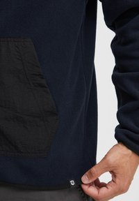Sudadera de forro polar en color azul marino con un bolsillo de nailon negro, tejido texturizado y una pequeña etiqueta de logo en contraste en el dobladillo.