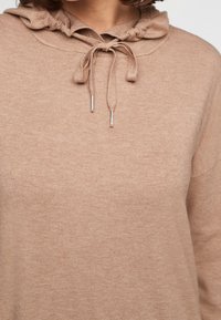 Sweat à capuche en maille beige avec un cordon de serrage au niveau du cou, porté par une personne partiellement visible des épaules au milieu de la poitrine.