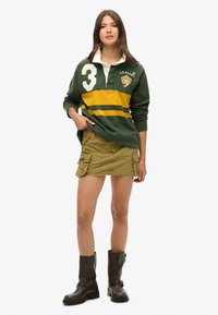 Grünes Rugby-Shirt mit weißem Kragen, gelben Streifen und der Nummer 3; olivgrüne Cargo-Rock; braune kniehohe Stiefel. Lässiges sportliches Outfit.