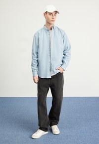 Chemise bleu clair à boutons, t-shirt gris en dessous, pantalon foncé, baskets blanches et casquette blanche, debout sur un sol bleu contre un fond uni.