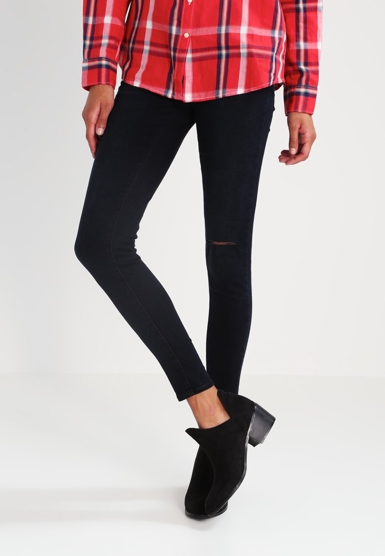 Jean skinny noir avec un genou déchiré, associé à une chemise à carreaux rouge boutonnée et des bottines noires, avec une finition en daim texturé.