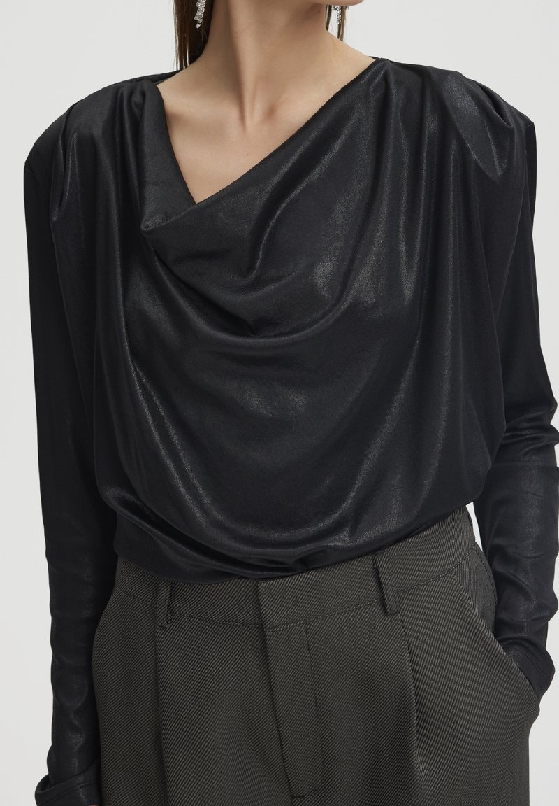Femme portant un chemisier noir brillant à manches longues drapé, rentré dans un pantalon taille haute gris foncé coupé sur mesure, les mains dans les poches.