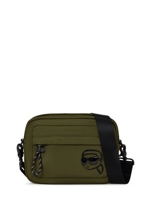 Olivově zelená obdélníková crossbody taška s černým nastavitelným popruhem a kapsou na zip, s černým stylizovaným logem obličeje se slunečními brýlemi.