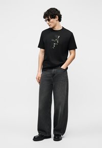 Νεαρός άνδρας που φοράει μαύρο T-shirt με σχέδιο, φαρδιά σκούρα γκρι τζιν, μαύρα παπούτσια και μαύρα γυαλιά ηλίου, στέκεται με το ένα χέρι στην τσέπη.