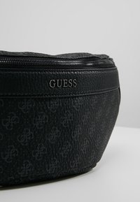 Pochette zippée noire texturée Guess avec motif de logo en relief et nom de marque métallique "GUESS" sur le panneau avant.