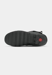 Semelle de chaussure en caoutchouc noir avec une surface texturée, présentant des rainures horizontales et un accent circulaire rouge, ainsi qu'un logo de marque.