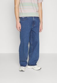 Jeans de mezclilla azul con un corte holgado, con costuras visibles y un logotipo de la marca en la cintura, combinados con una camisa de rayas de manga corta.