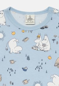 Ljusblå bomullströja med Moomin-karaktärer i olika lekfulla scener, inklusive träd, muggar och frukter, med rund halsringning.