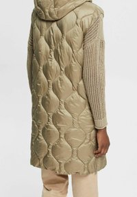 Długa pikowana kamizelka w oliwkowej zieleni; posiada dzianinowe rękawy, kaptur oraz boczny zamek błyskawiczny do zamykania.