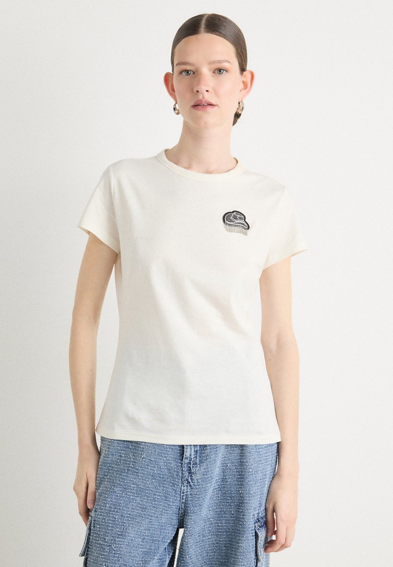 Cream cotton t-shirt with short sleeves, round neckline, and black embroidered design on the chest. Paired with blue textured denim shorts.