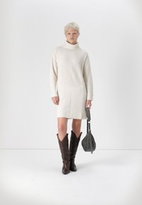 Πλεκτό κρεμ φόρεμα με ψηλό γιακά, ραβδωτό τελείωμα και υφασματική ψηφιά. Φορώντας με καφέ cowboy μπότες και γκρι τσάντα ώμου.