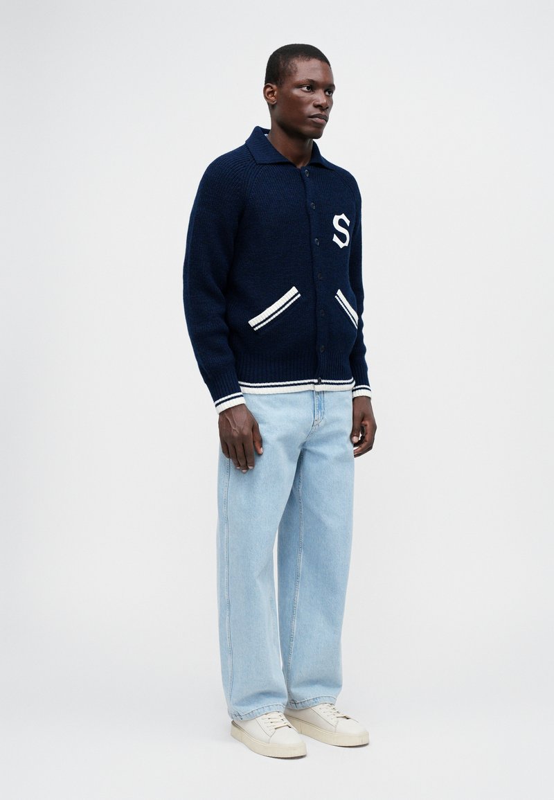 Marineblauwe gebreide cardigan met witte accenten en een "S"-logo, gecombineerd met lichtblauwe wijde jeans en crèmekleurige sneakers op een effen achtergrond.